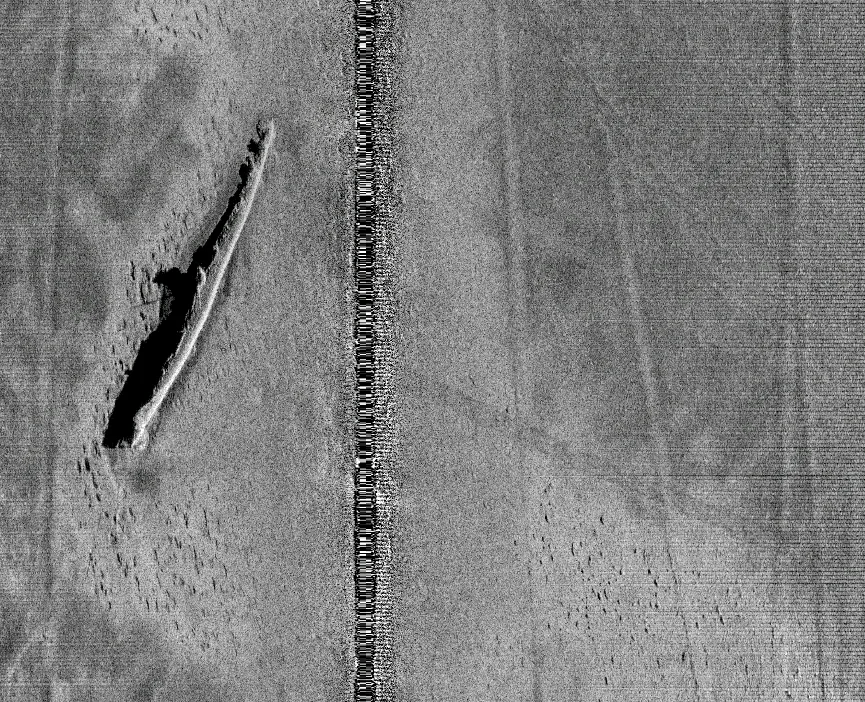 Gray side-scan sonar image of a sunken submarine lying on a sandy bottom, with a long black shadow beside it and a noisy line down the center of the image